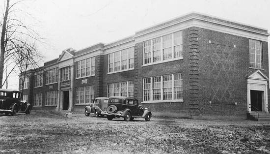Black and white photograph of the old Fairfax High School taken in 1935. The building is two stories tall with six large banks of windows on both floors. The main entrance is ornately designed with columns. Early 1930s era cars are parked in front of the building.  
