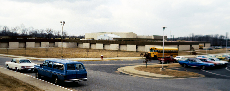 Undated color photograph of the new Fairfax High School taken from the hillside along Old Lee Highway. In the foreground are several cars and a school bus which, by their makes and models, appear to date this photo to the late 1970s or early 1980s. 