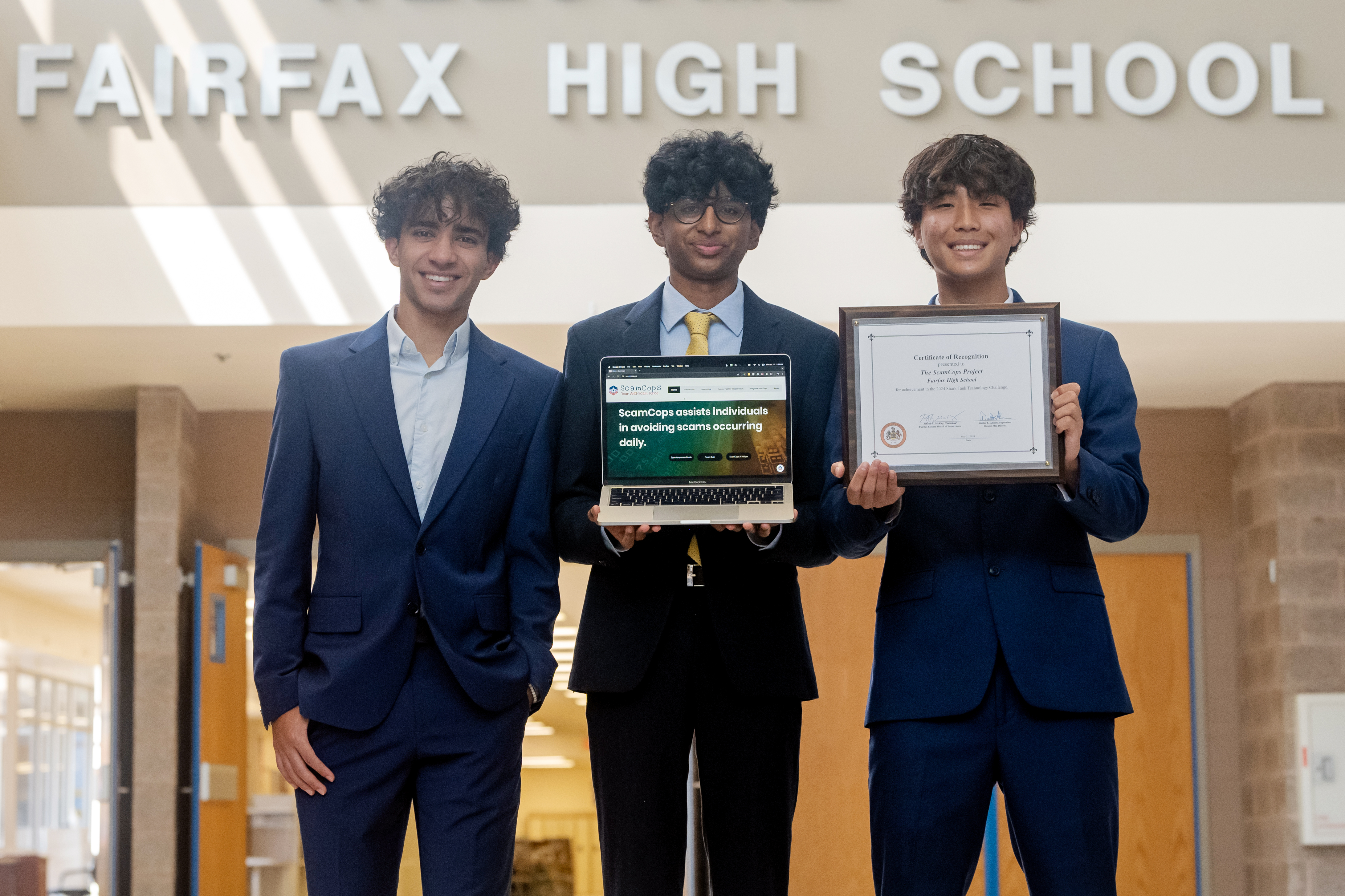 Fairfax High School students Yousif Al Atbi, Advik Atyam, and David Nam stand, smiling, under a large sign that reads: "FAIRFAX HIGH SCHOOL." Advik, at center, holds a laptop logged in to the ScamCops website. At right, David holds an award plaque the students received from Fairfax County.